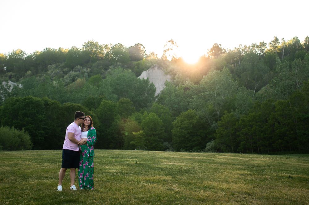 Couple embracing in a sunlit green field with trees and golden sunset in the background.