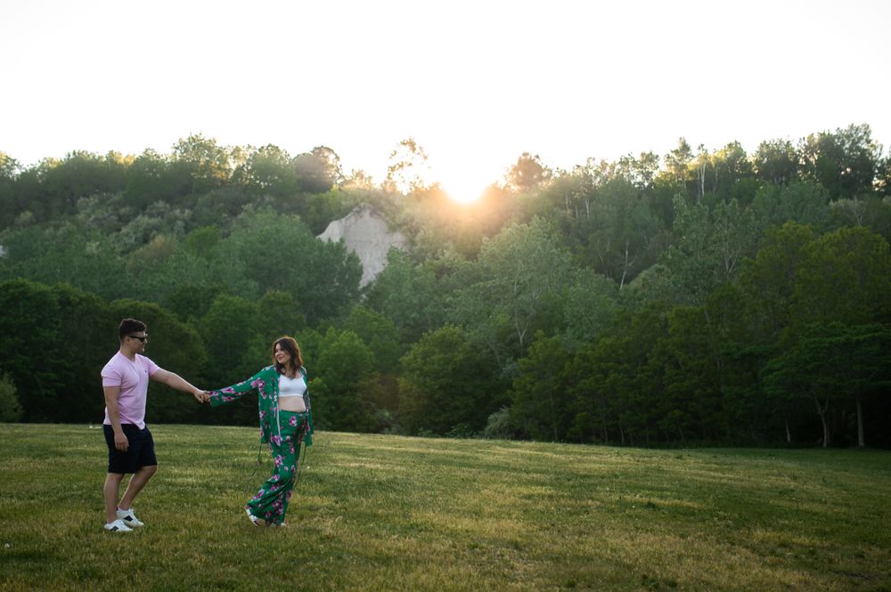 Couple holding hands dancing in a green meadow at golden sunset with lush forest trees in background.