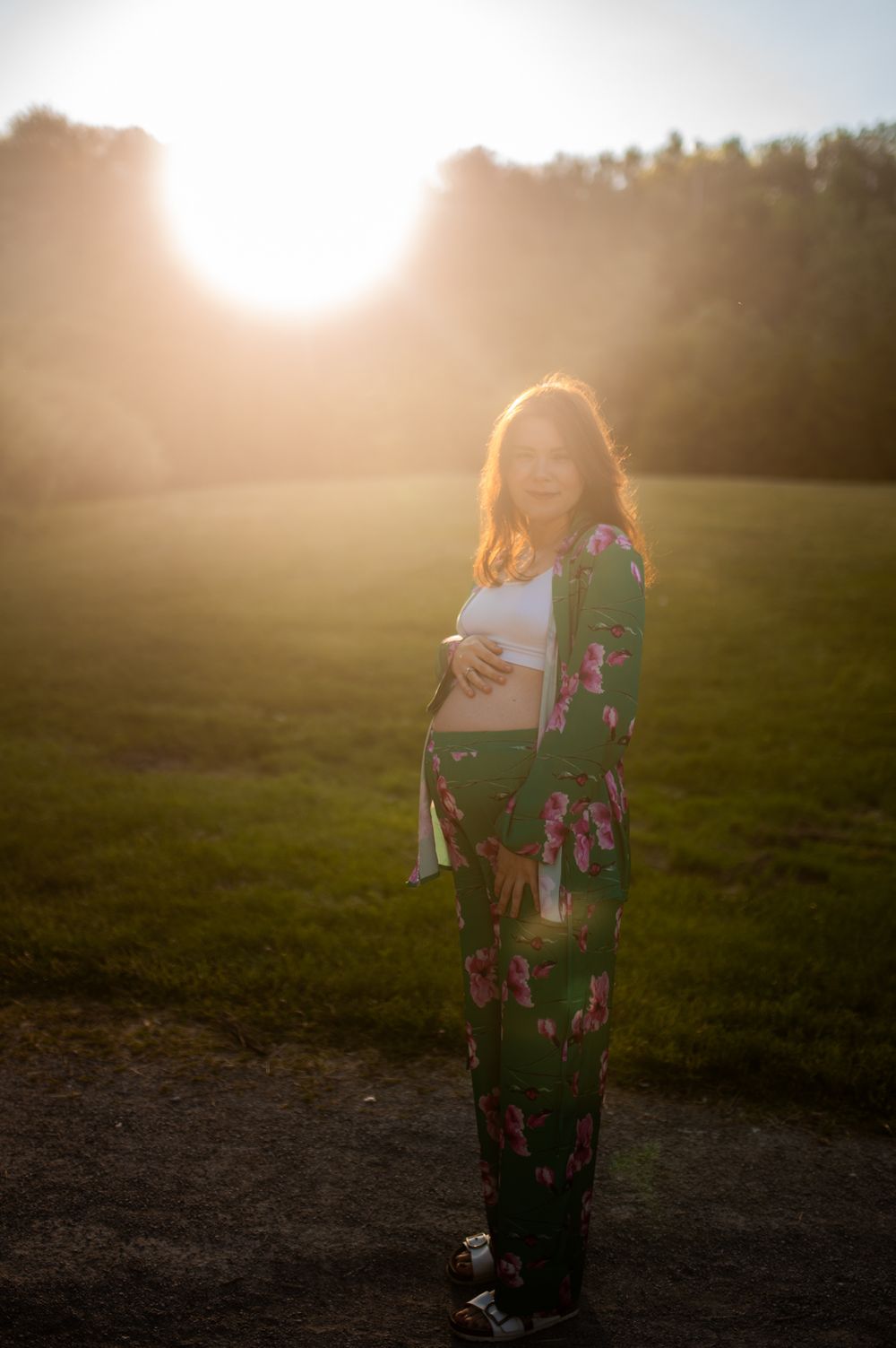 Pregnant woman in floral kimono cradling baby bump in a sunlit green field during golden hour.