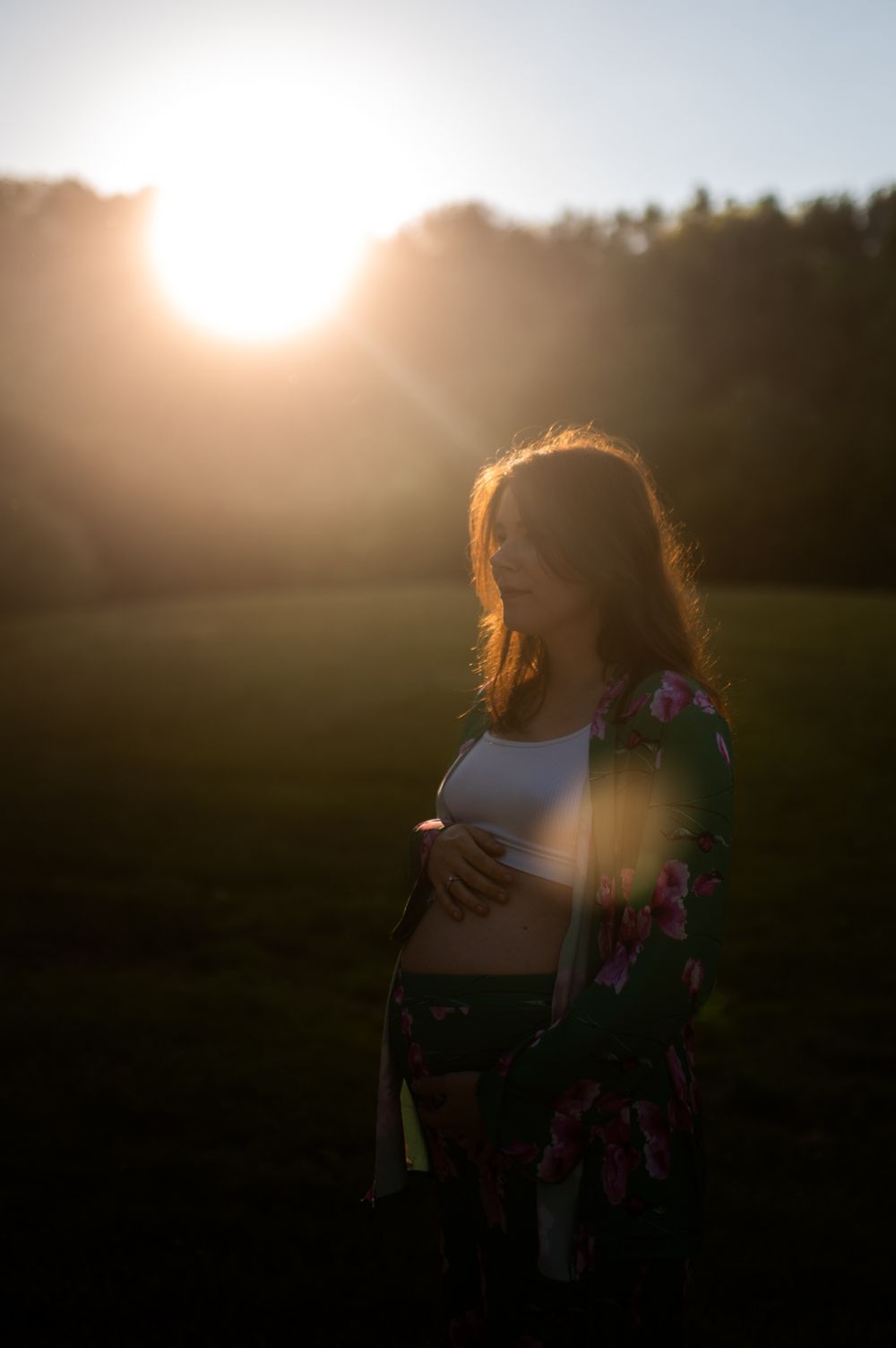 Pregnant woman cradling baby bump in golden sunset light wearing floral kimono in a green field.