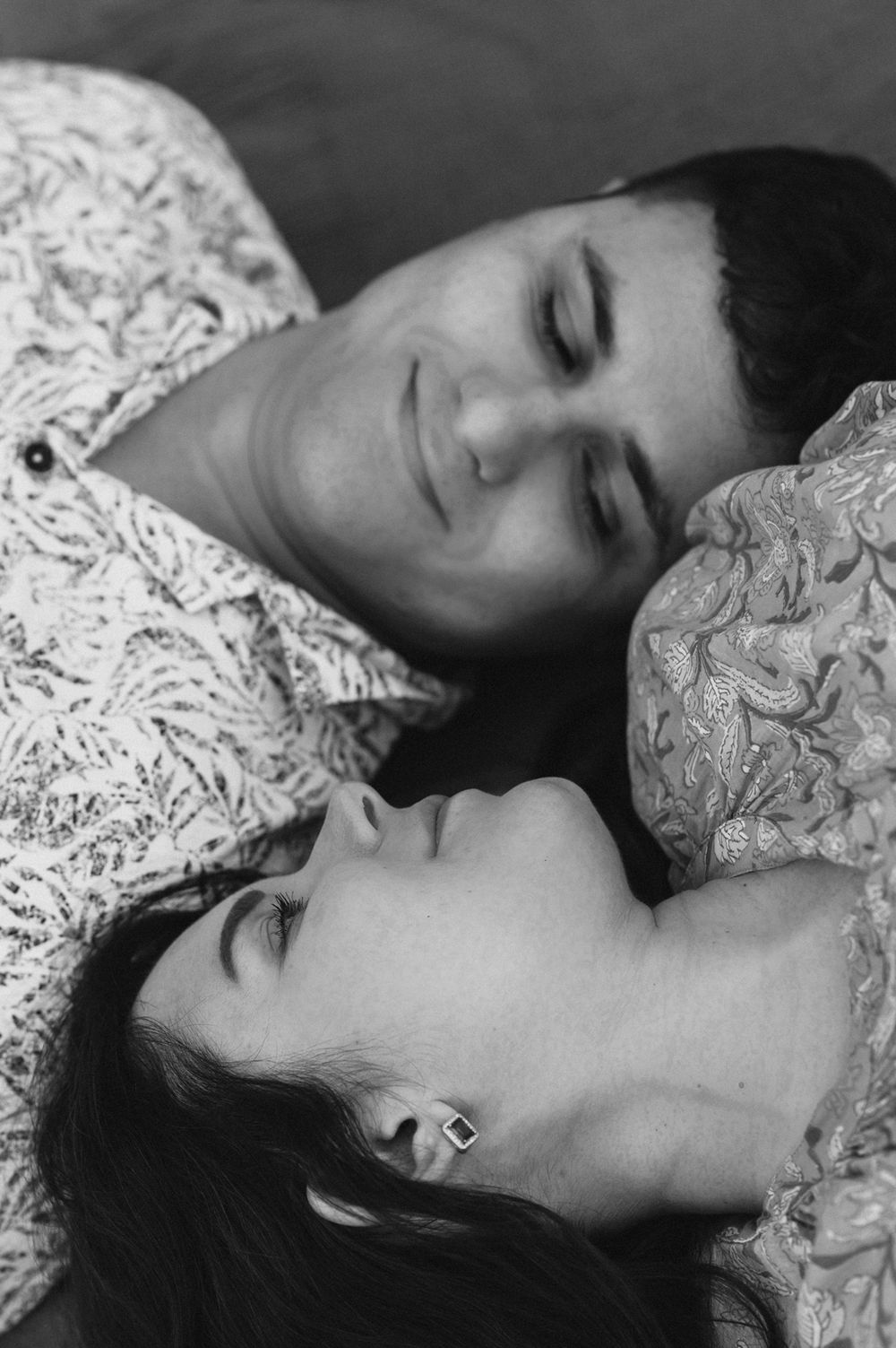 Black and white photo of a couple lying down together with eyes closed, wearing patterned clothing.