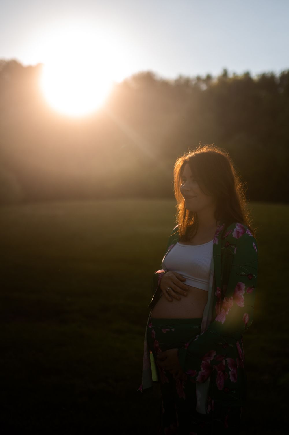 Pregnant woman gently holding belly at sunset in a green field, wearing a floral kimono outdoors.