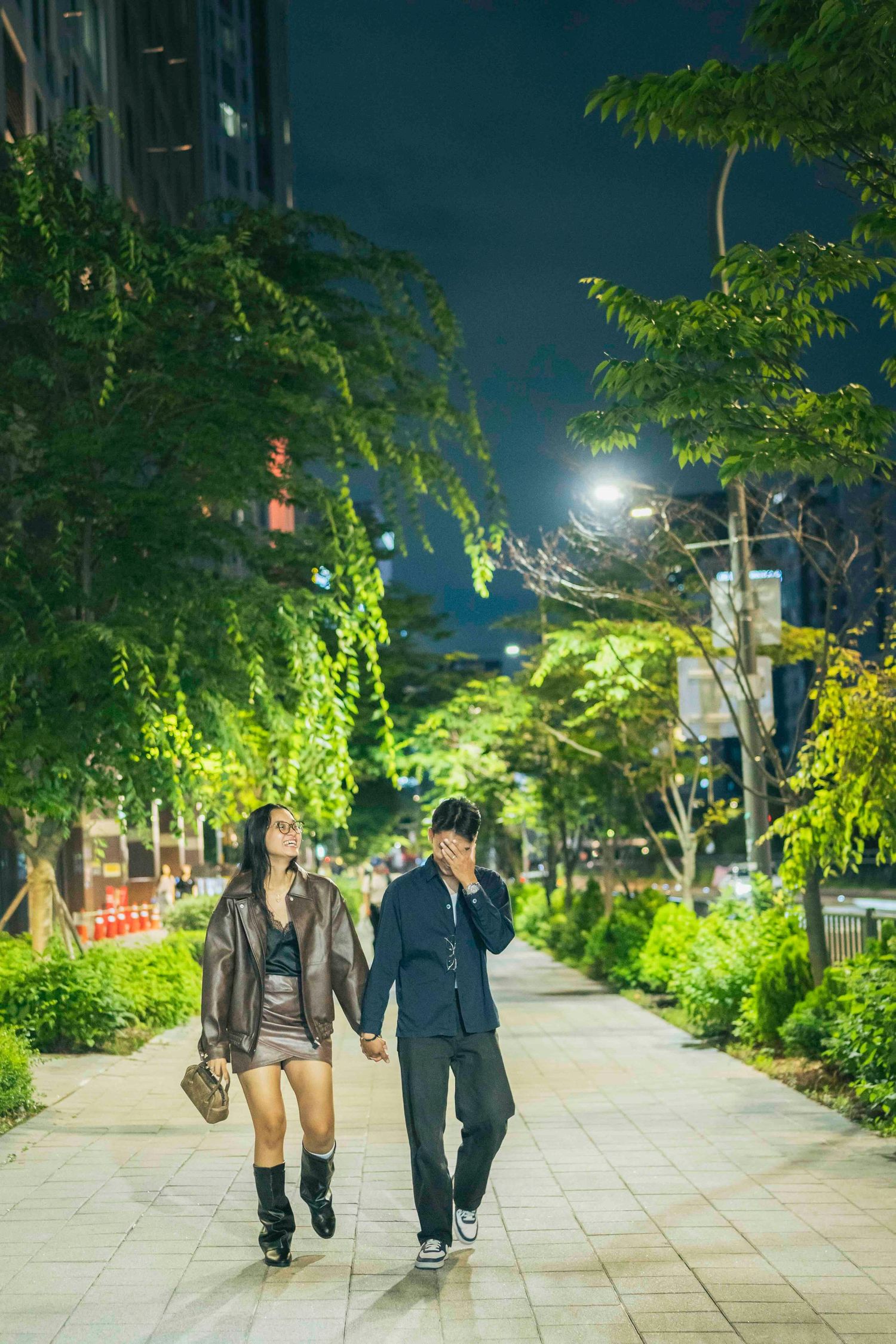 Couple walking hand in hand on a tree-lined city sidewalk at night, illuminated by street lights.