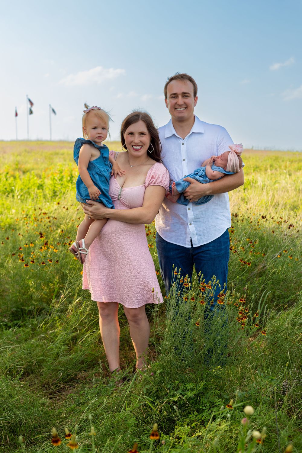 Happy family of four posing in a wildflower field, parents holding two young children under a blue sky.