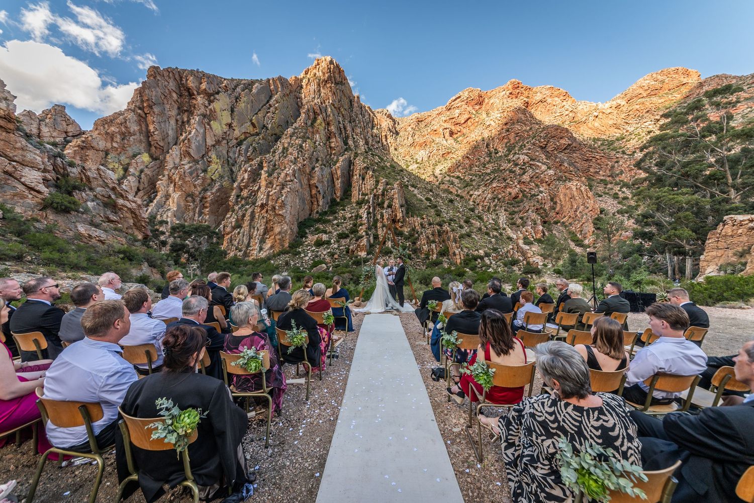 Outdoor wedding ceremony in Red Rock Canyon with guests seated along aisle, dramatic rocky mountains backdrop under blue sky.