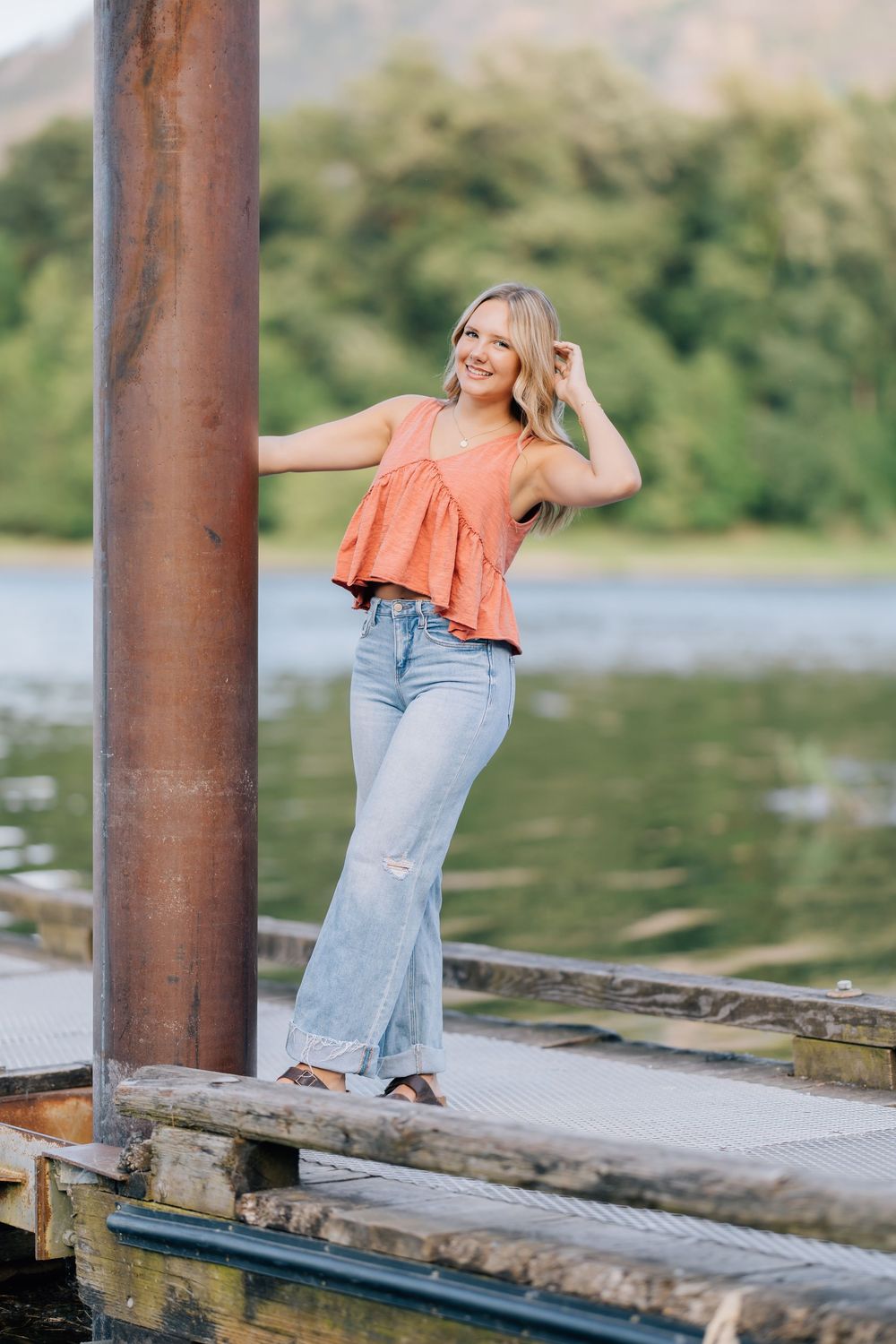 Smiling blonde woman in orange top and flared jeans posing on a wooden dock by a lake.
