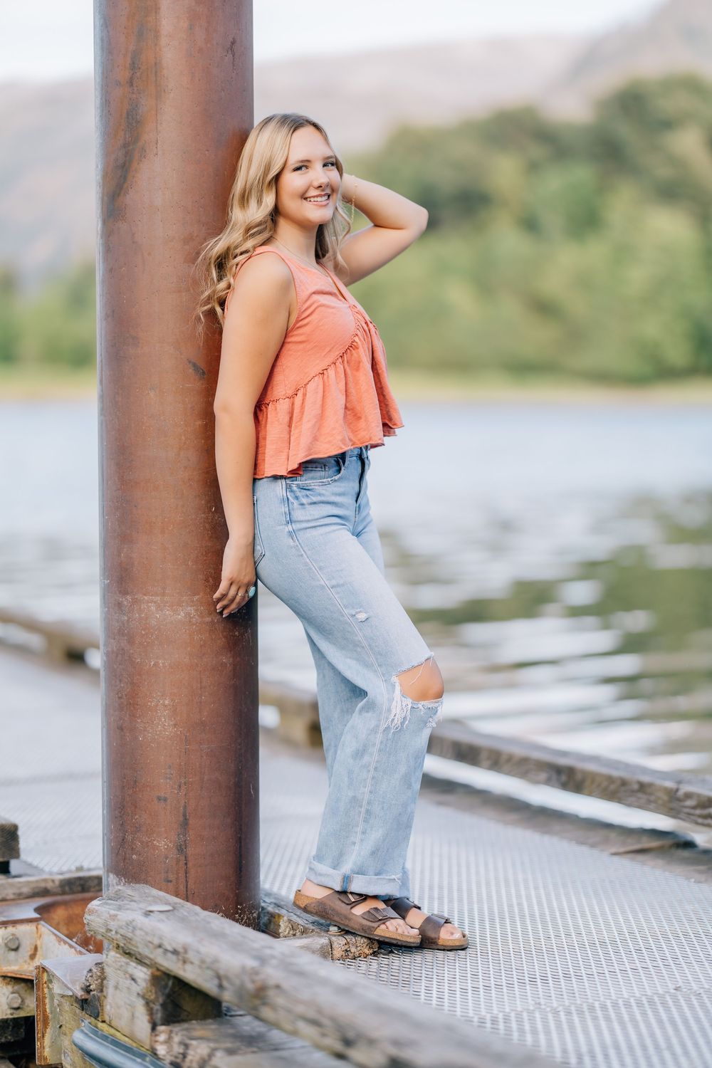 Blonde woman in coral top and ripped jeans posing on a dock by a lake with mountains in the background.