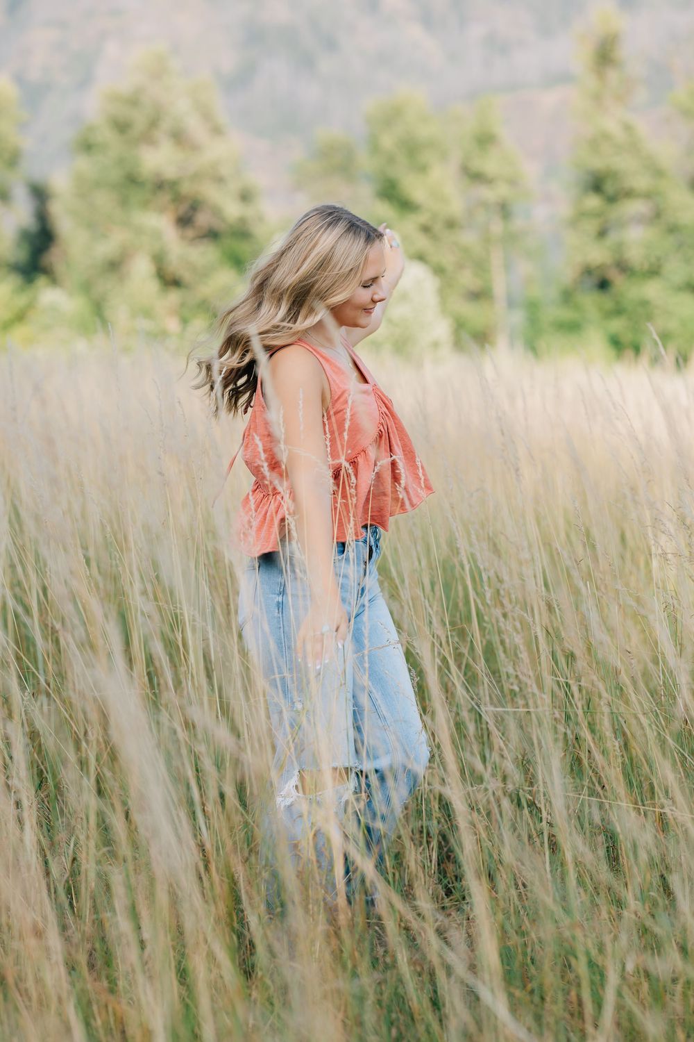 Blonde woman in coral top and jeans walking through tall golden grass field with mountains behind.