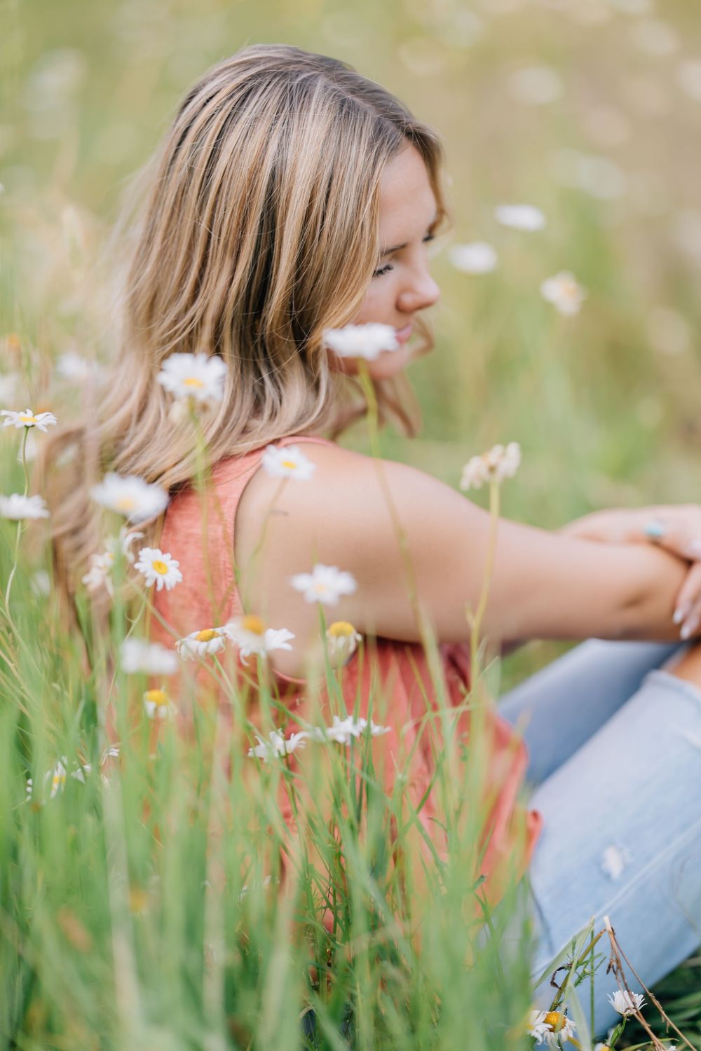 Blonde woman sitting in a field of white daisies wearing an orange tank top and jeans.