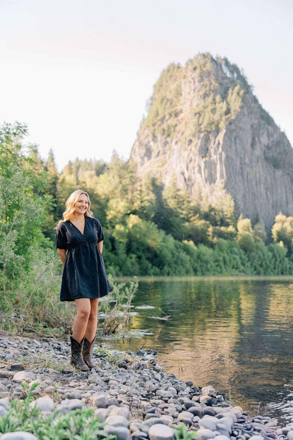 Blonde woman in dark dress and cowboy boots standing on rocky riverbank with dramatic cliff backdrop.