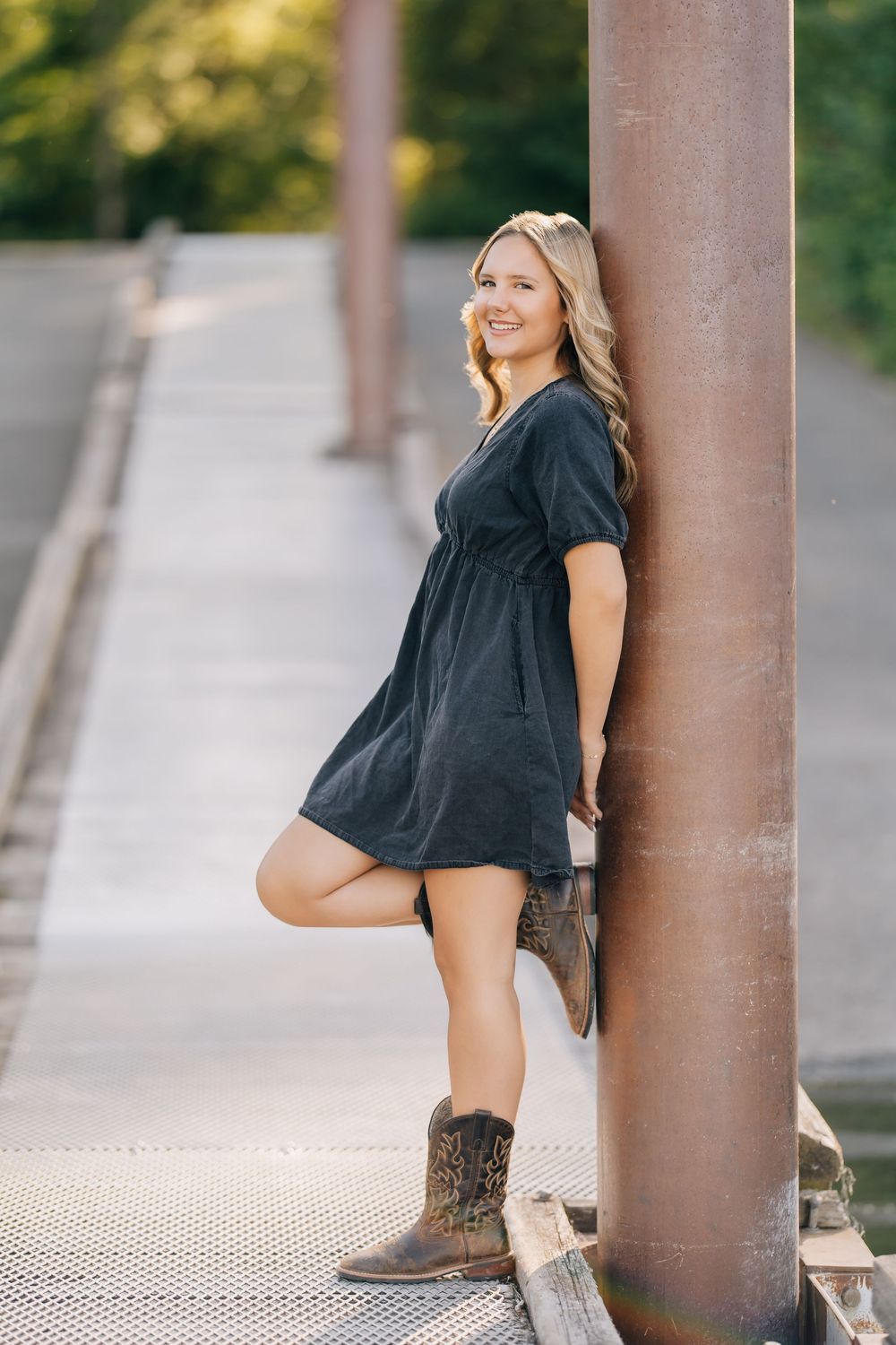 Smiling blonde woman in dark dress and cowboy boots leaning against rustic metal pillar on bridge.