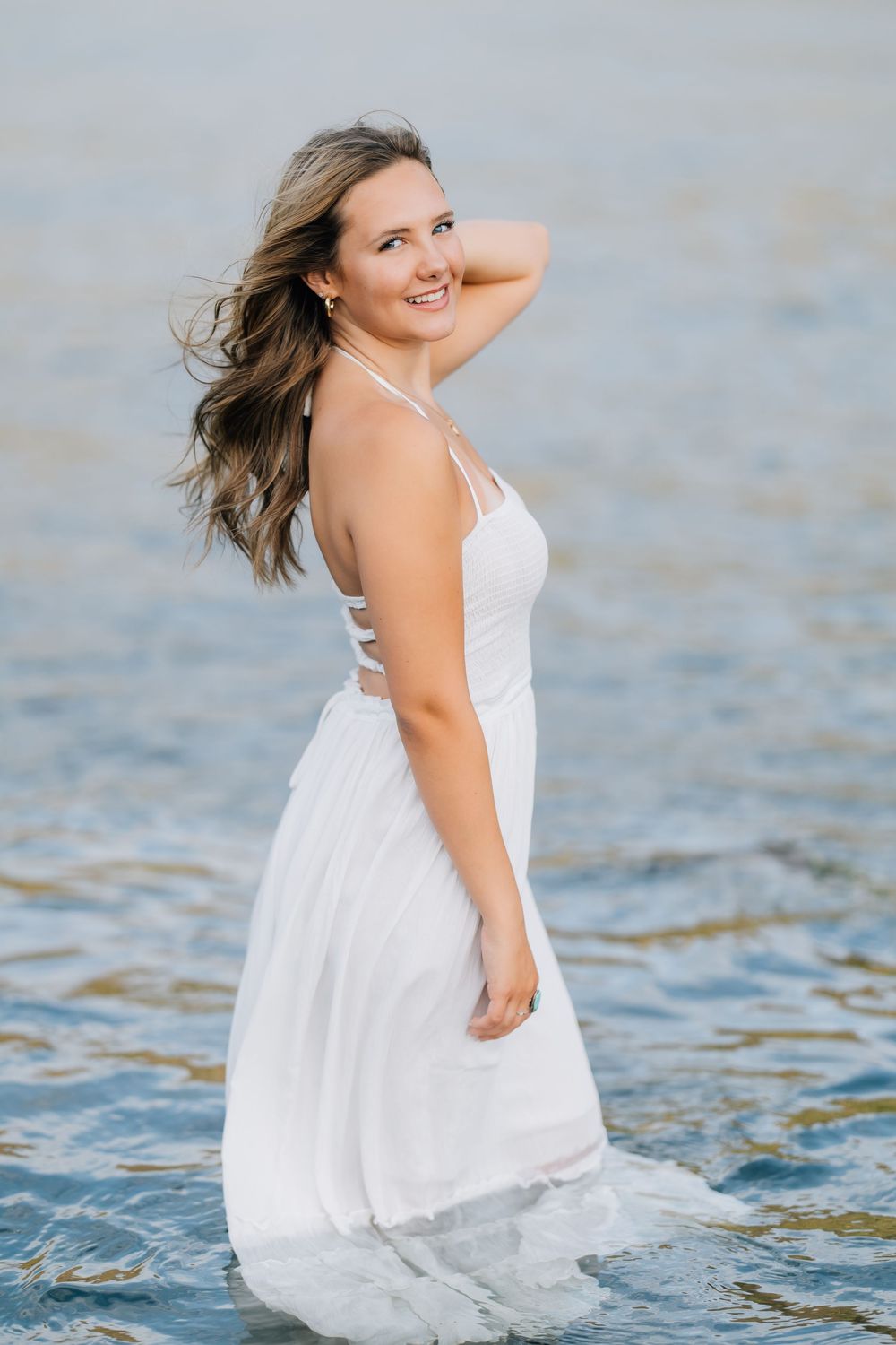 Smiling young woman in white halter dress standing in shallow water with wind-blown hair.