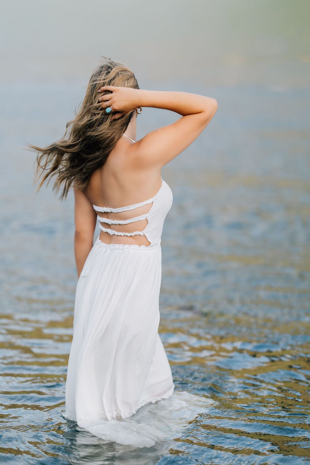 Young woman in backless white dress standing in shallow water with hand in flowing hair.