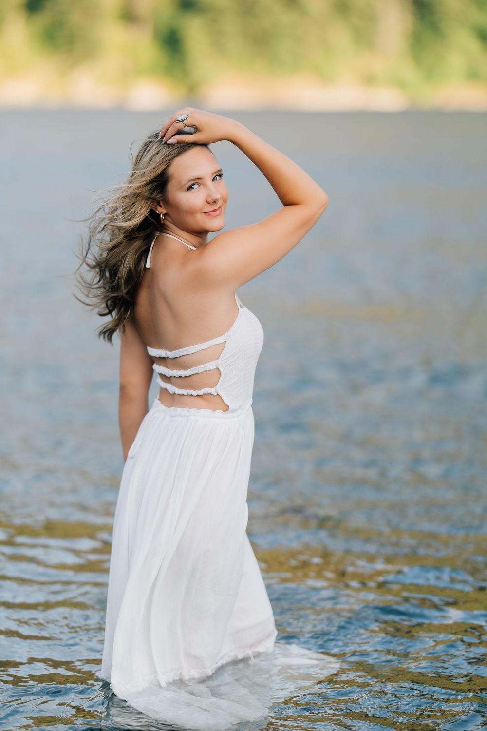 Woman in white backless dress standing in water with wind-blown hair, smiling over her shoulder.