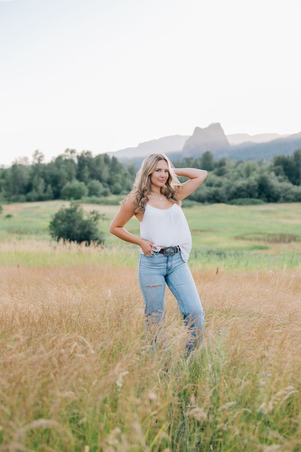 Blonde woman in white tank and ripped jeans standing in a golden meadow with mountains and trees behind her.