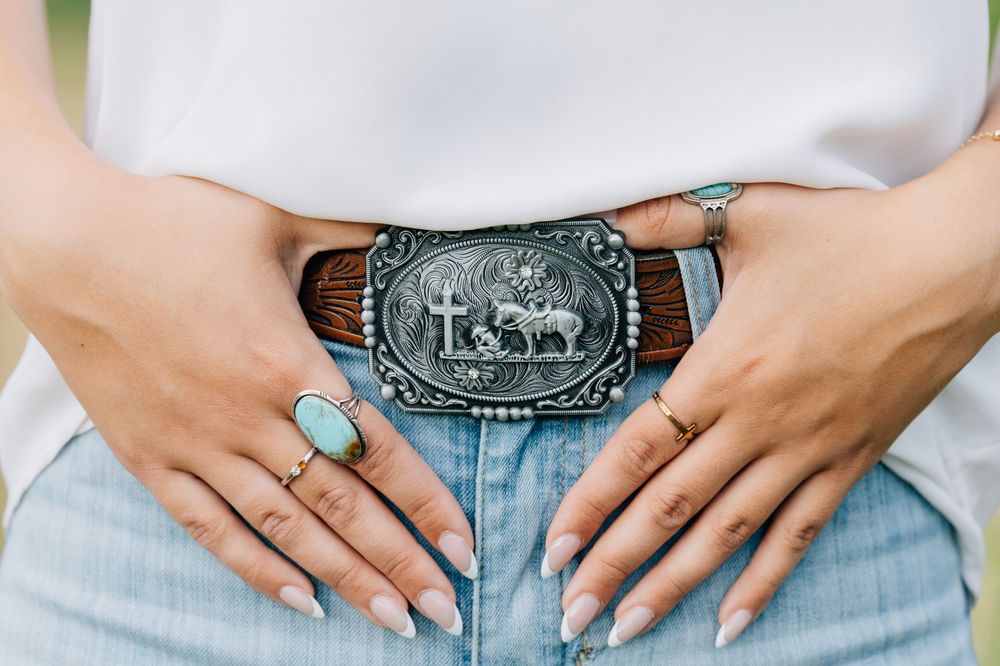 Woman wearing jeans with a silver western rodeo belt buckle, turquoise ring, and white manicured nails.