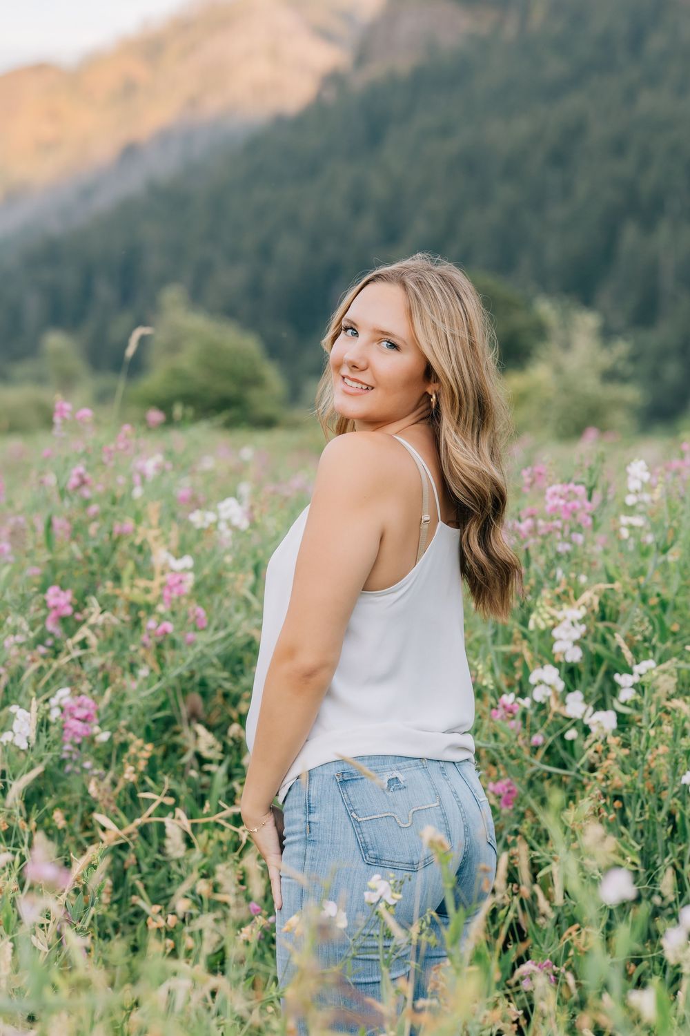 Smiling blonde woman in white tank and jeans posing in vibrant wildflower meadow with mountain views.