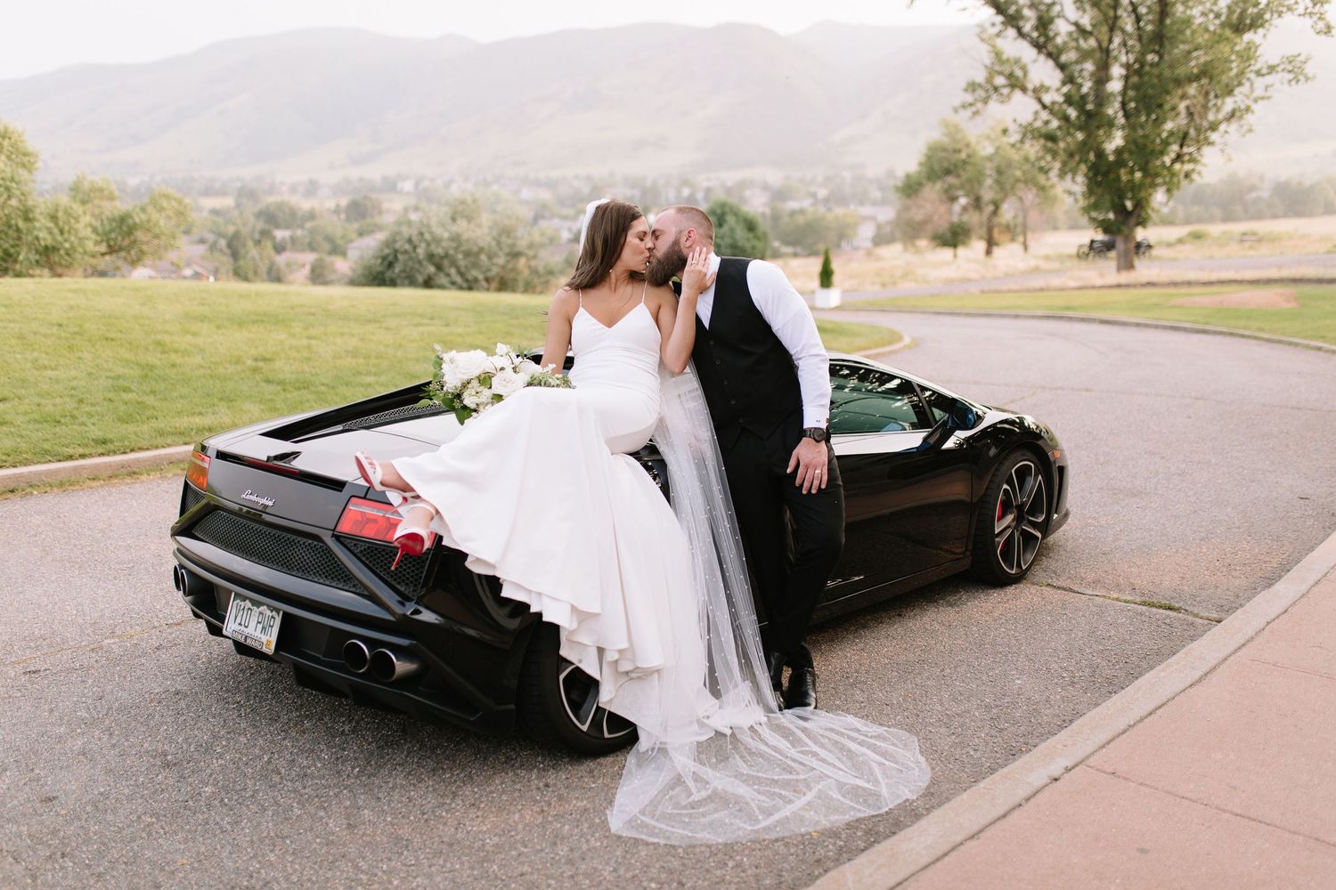 Bride and groom sharing a kiss leaning against a black Lamborghini convertible with mountain views backdrop.