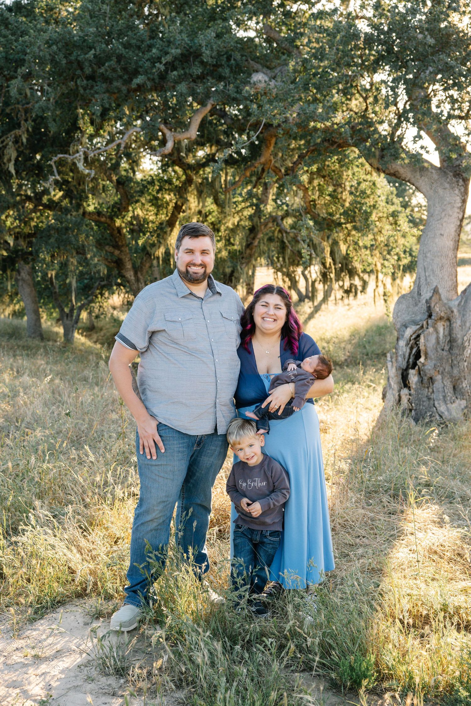 Family of four pose mom holding newborn baby and toddler standing below in grass and oak trees during sunset, golden hour photo