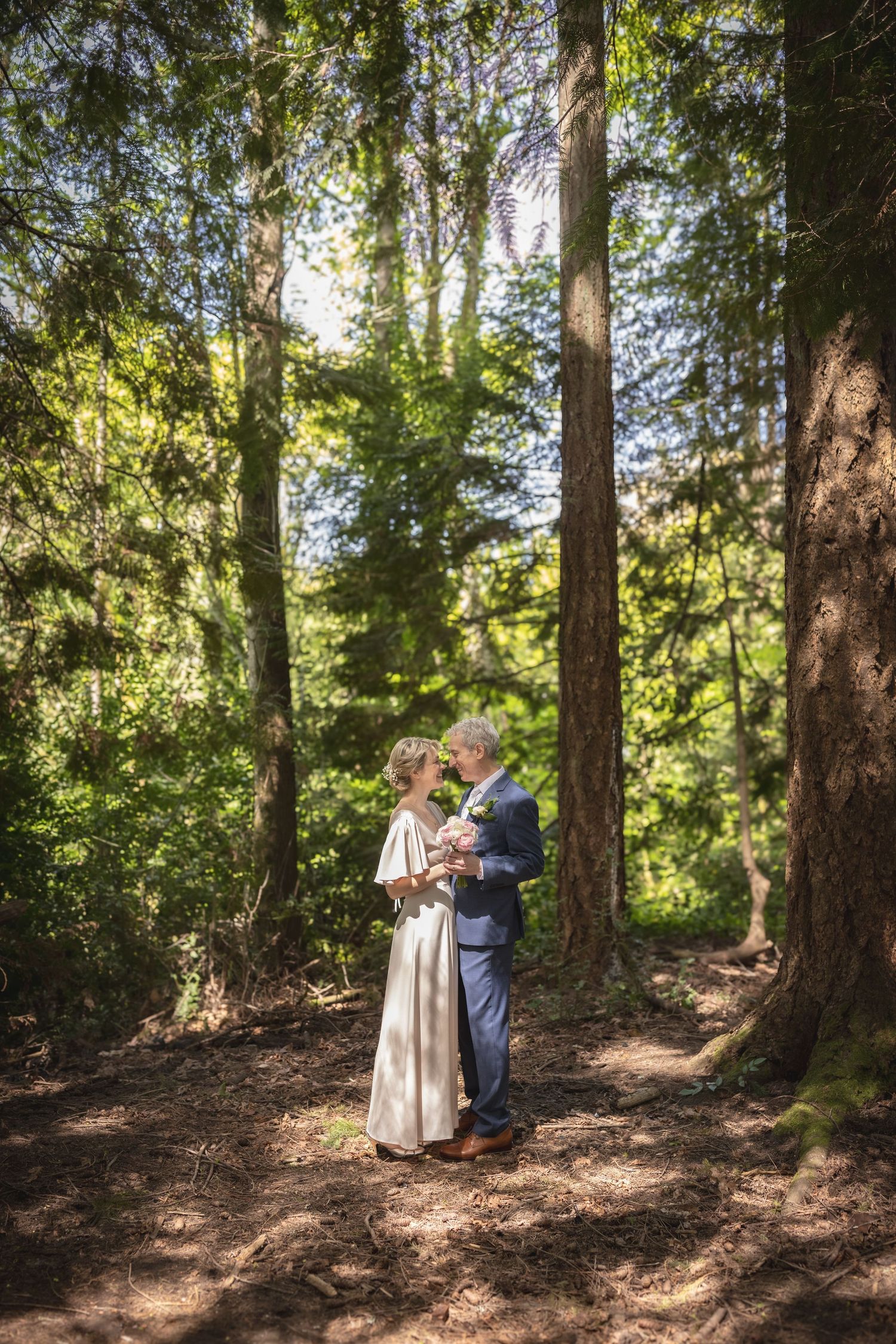 Bride and groom share a romantic embrace on a sunlit forest path surrounded by tall evergreen trees.