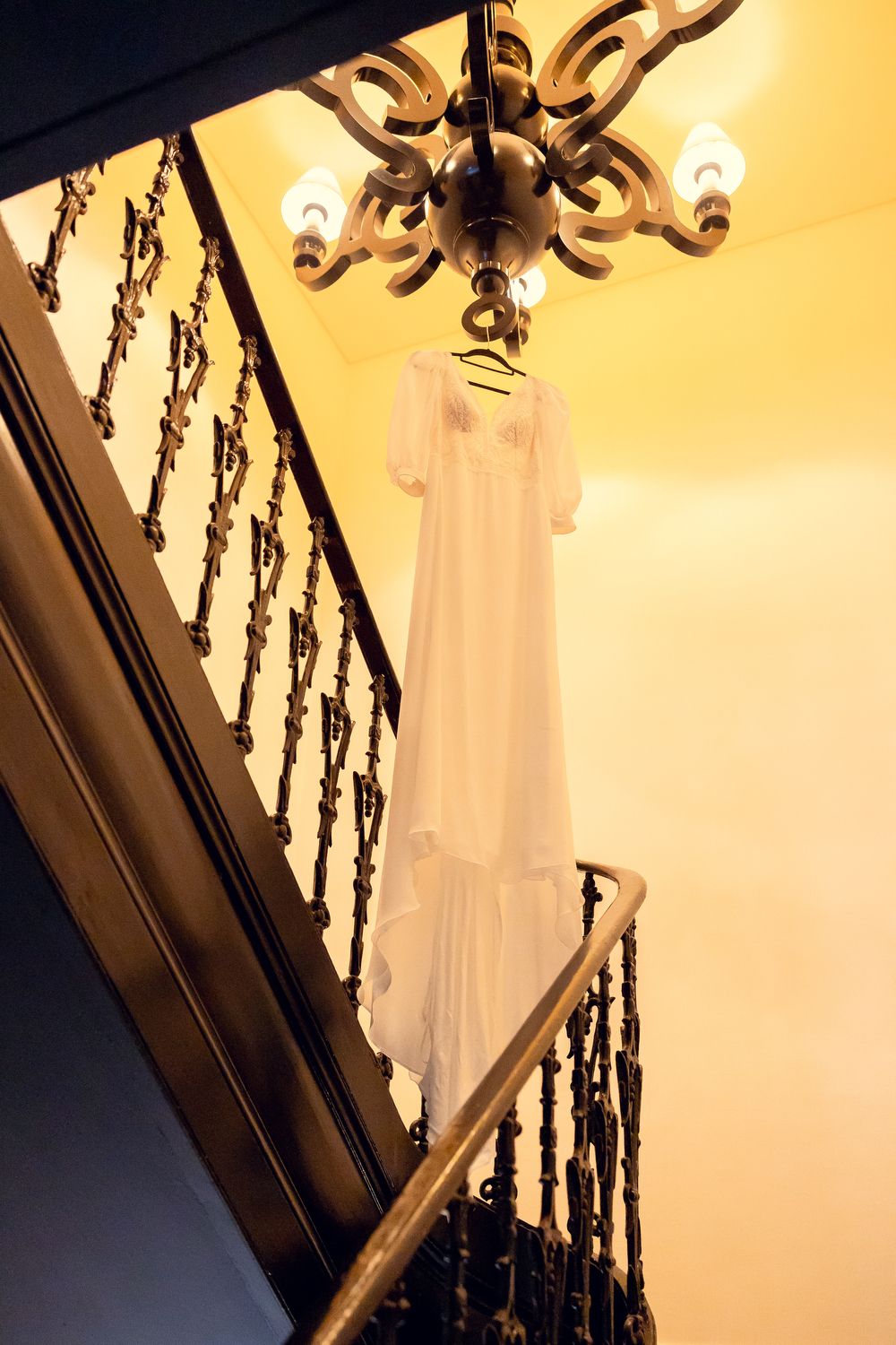 White wedding dress hanging from ornate chandelier over elegant staircase with wrought iron railings in warm light.