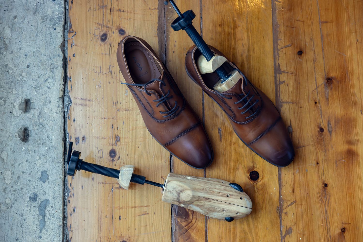Brown leather oxford shoes with wooden shoe trees on a rustic hardwood floor.