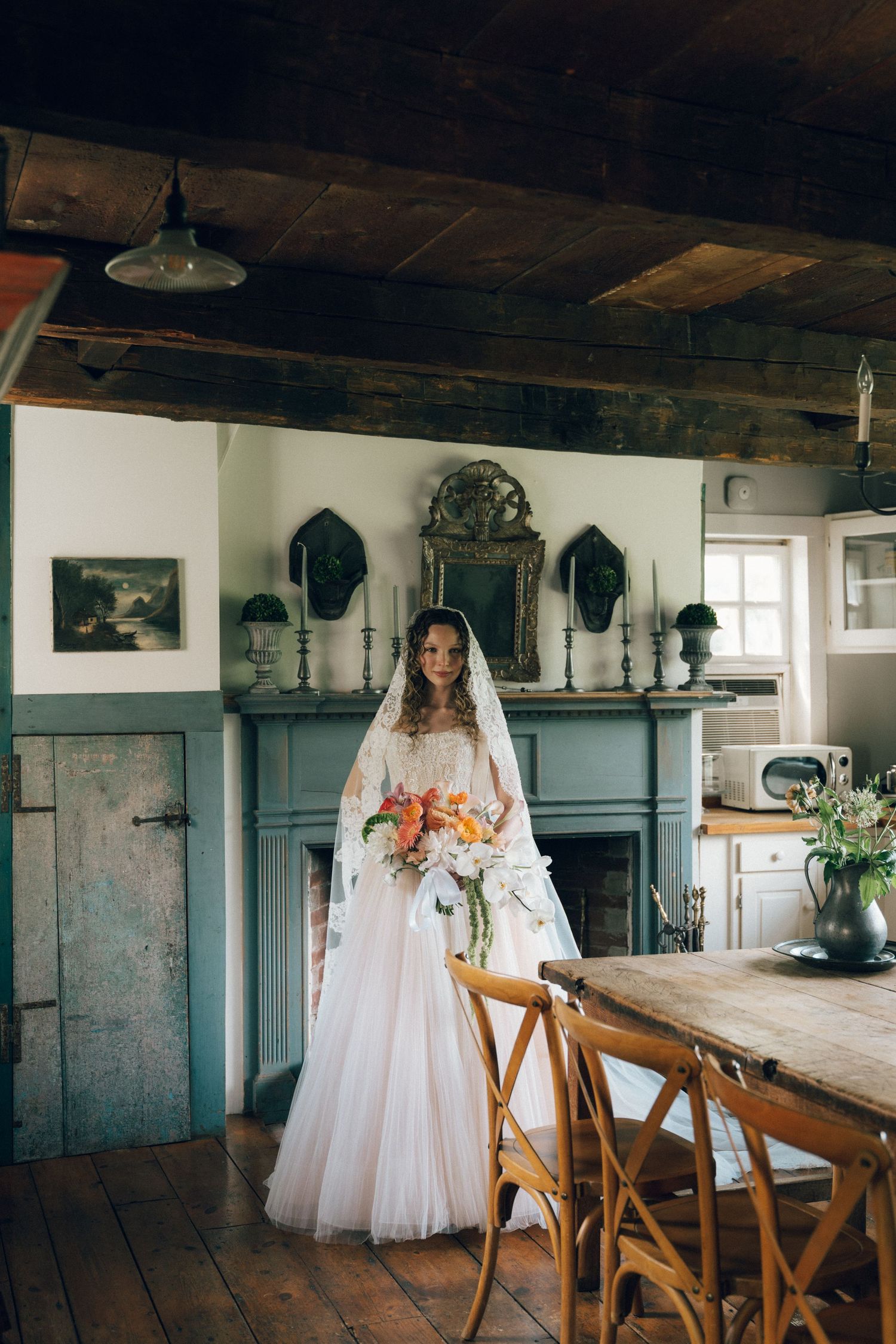 Bride in white gown and veil holding floral bouquet stands in rustic farmhouse dining room with stone fireplace.