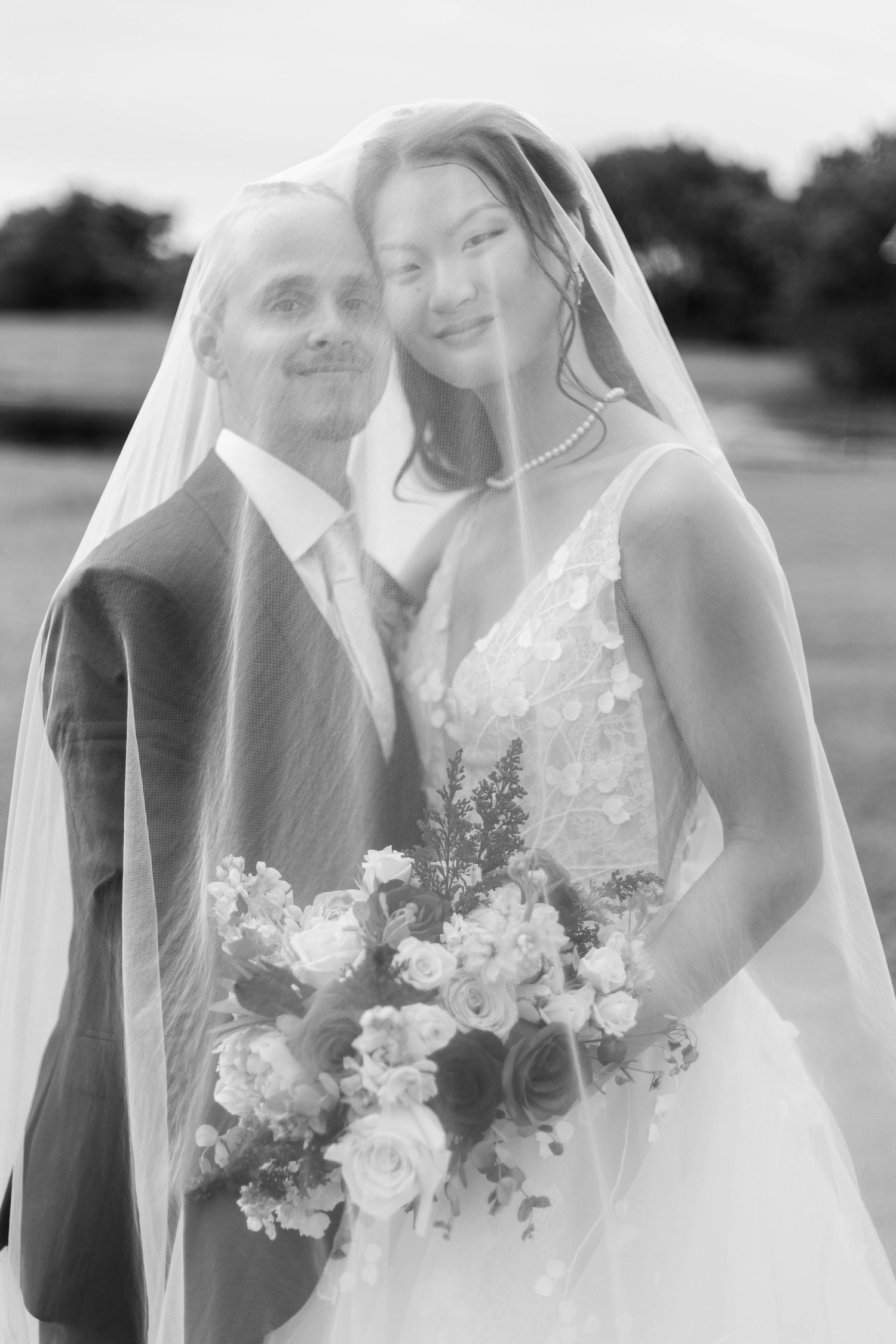 Black and white wedding photo of bride and groom under veil, bride holding bouquet of roses outdoors.