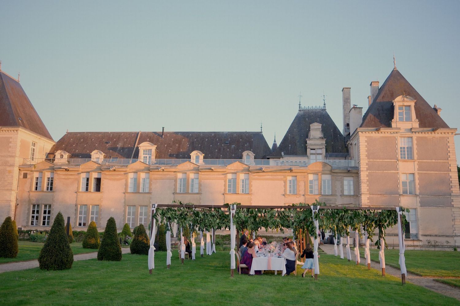 Wedding reception dinner set under floral pergola on lawn of grand French chateau at dusk.