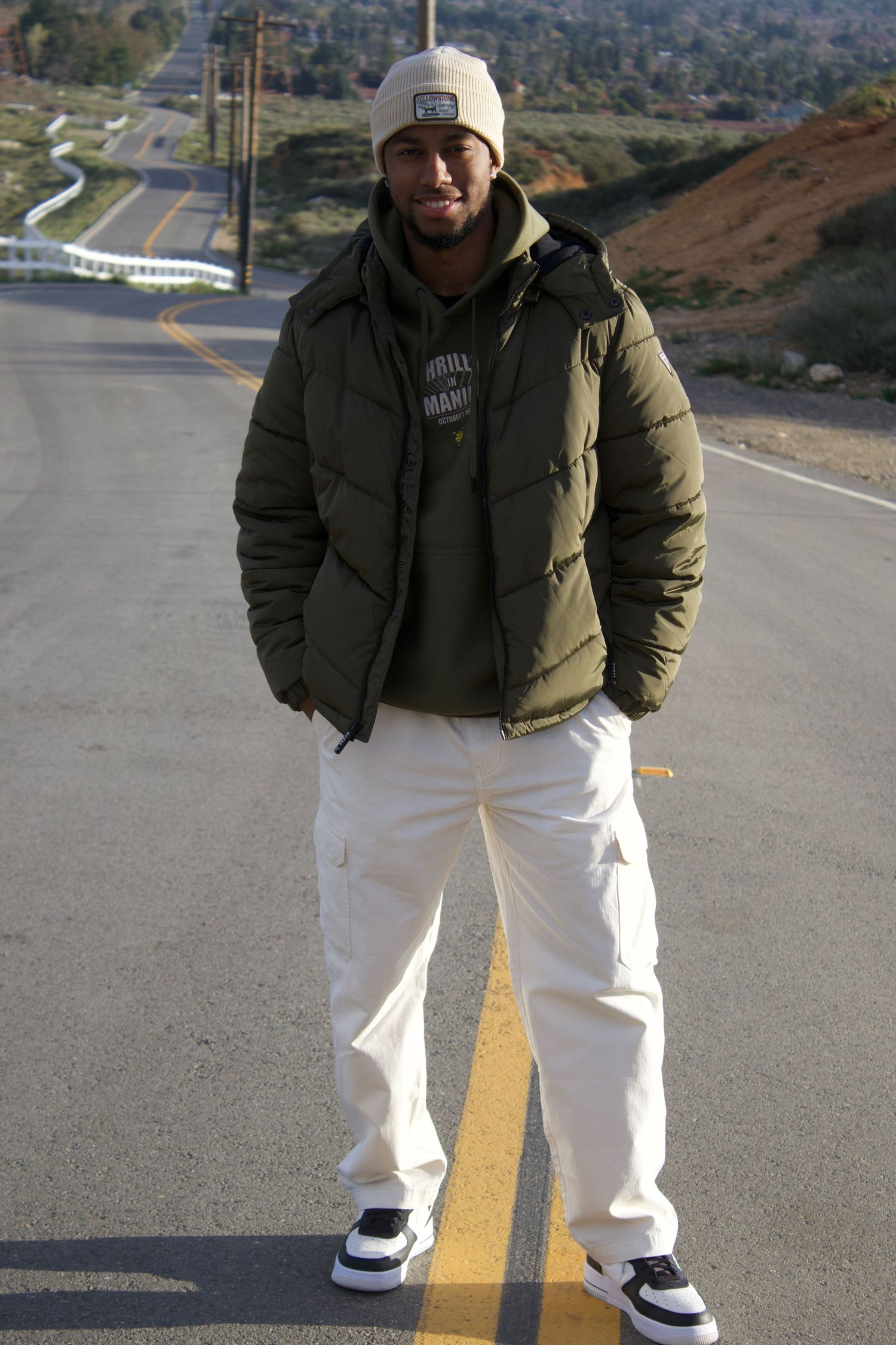 Man in olive puffer jacket and beanie stands on hilltop road with panoramic city valley view.