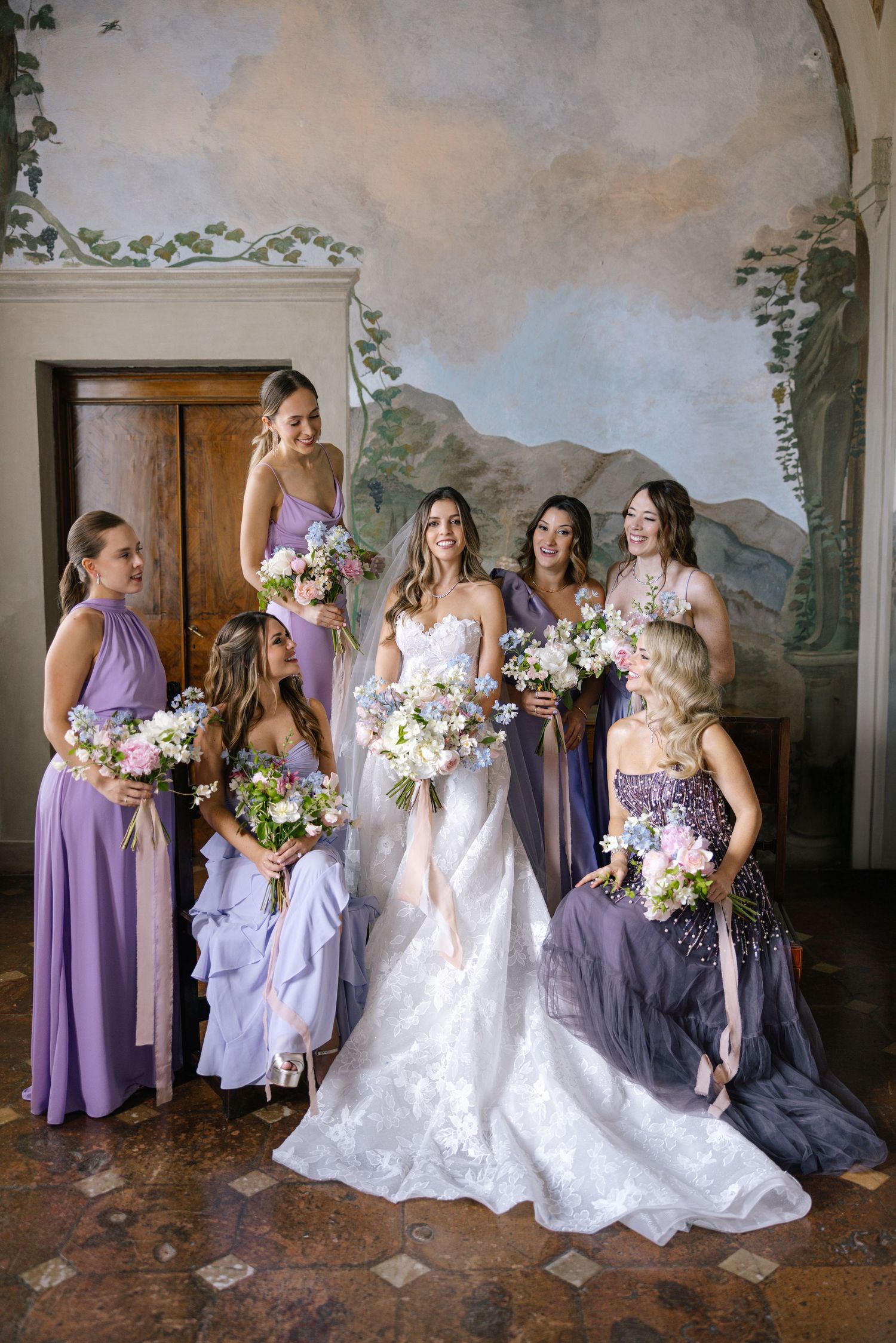 A couple walks down an outdoor aisle lined with flowers and cypress trees leading to an elegant villa entrance.
