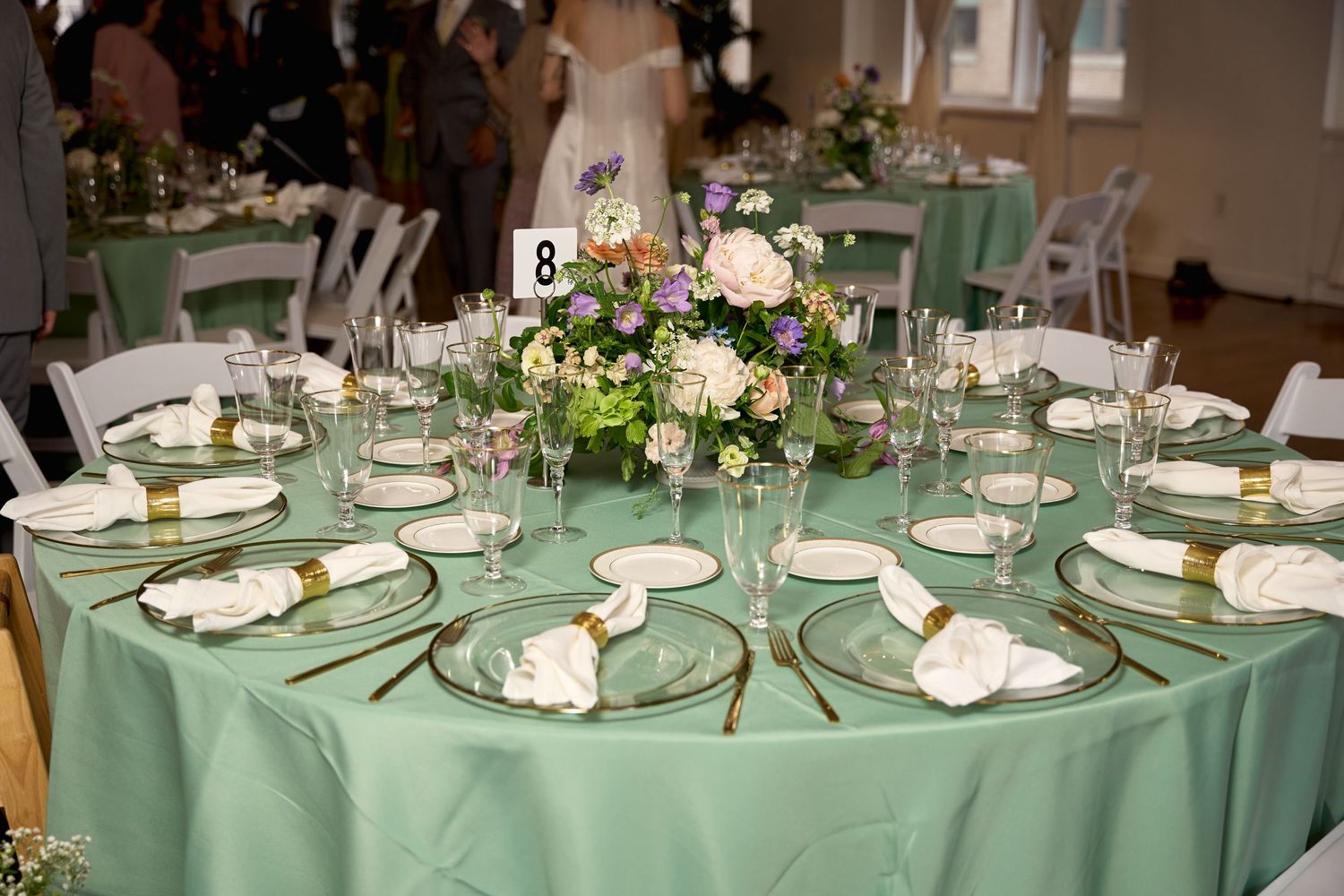 Elegant wedding reception table with mint green tablecloth, gold-rimmed plates, crystal glasses, and a floral centerpiece.