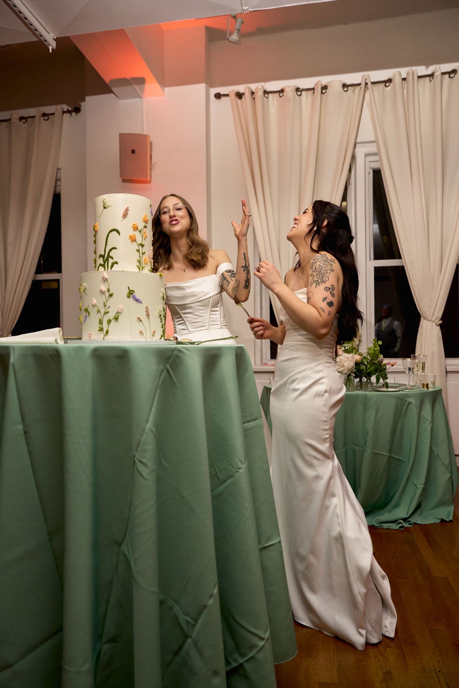Two brides in white gowns laugh joyfully beside a floral wedding cake on a green tablecloth at their reception.
