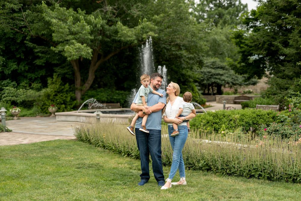 Family of four poses near a park fountain, parents holding two young children on a sunny day.
