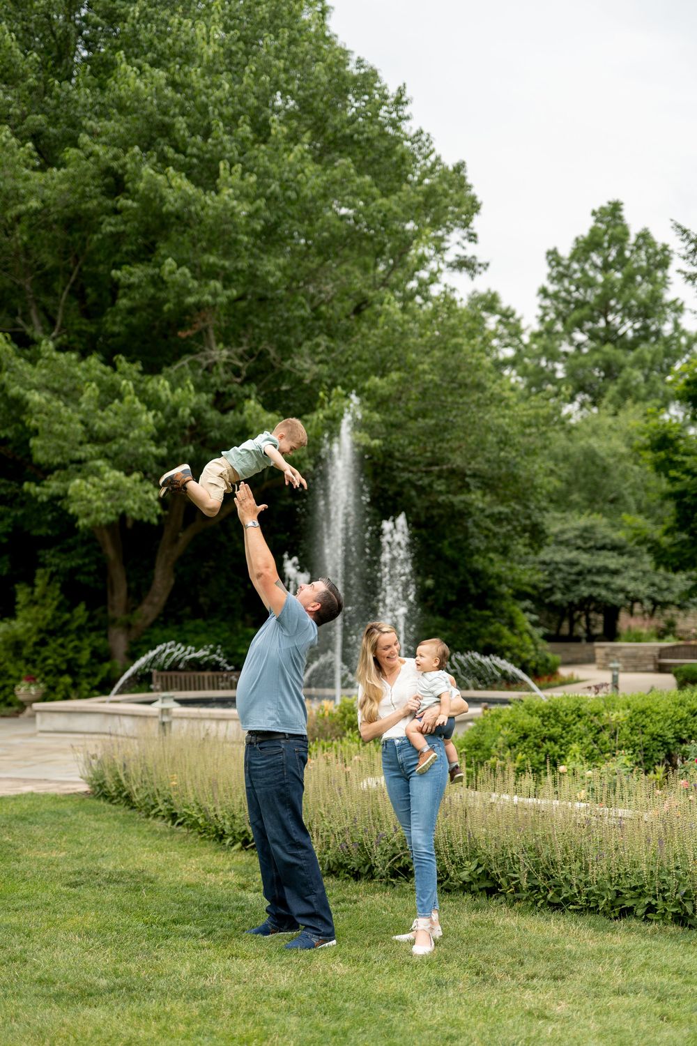 Father tosses toddler in the air while mother holds baby near a lush park fountain.