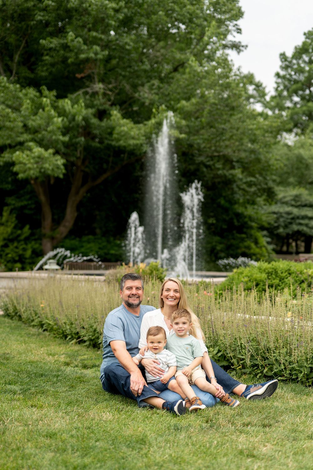 Happy family of four sits on green grass smiling, with a tall park fountain behind them.