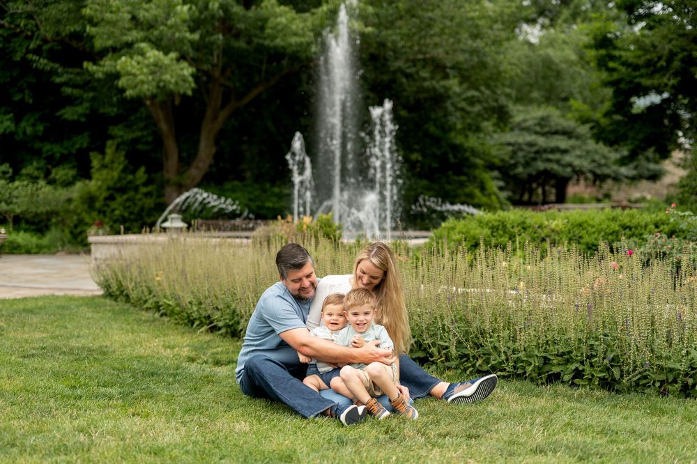 Family of three sitting on grass by a fountain in a lush green park on a sunny day.