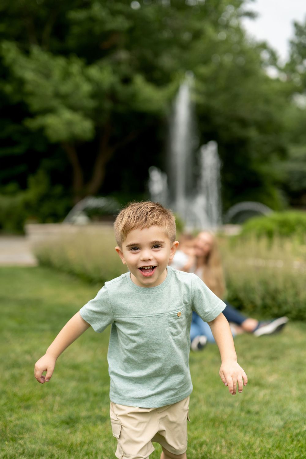 Excited young boy runs toward camera on green grass with a park fountain in the background.