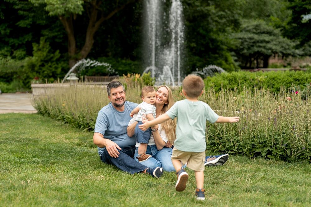 Family sitting on grass near fountain in park, parents smiling with toddler as young boy runs toward them.