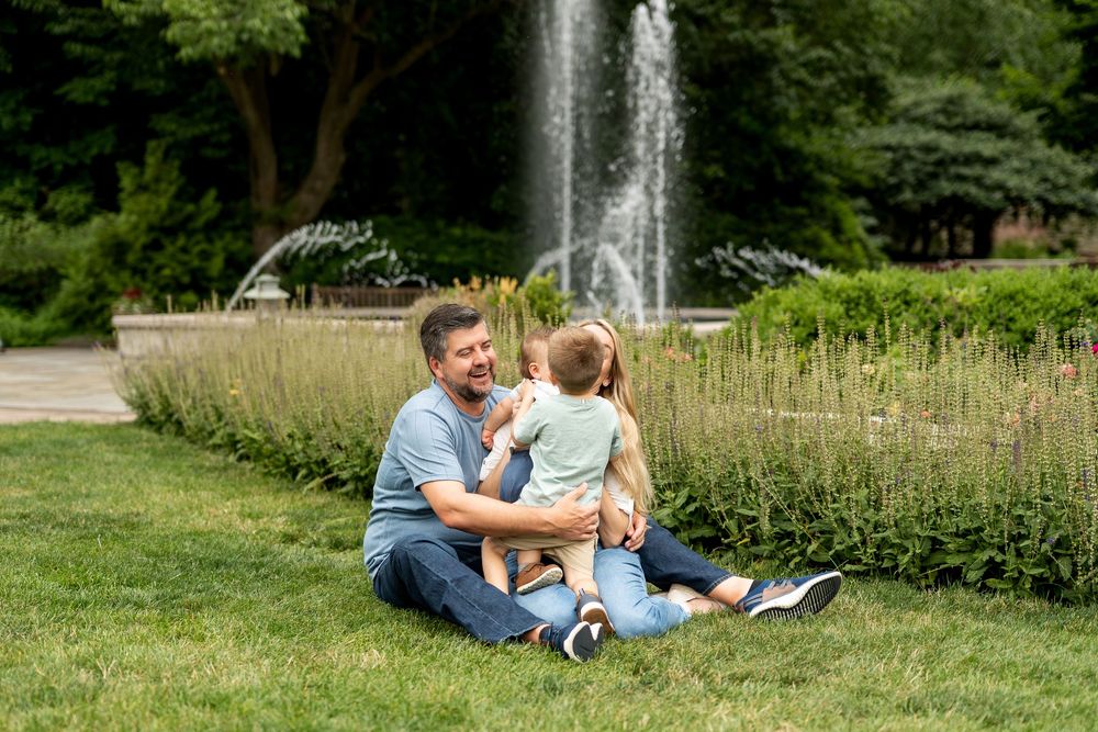 Father sitting on grass with toddler son in front of garden fountain surrounded by greenery.
