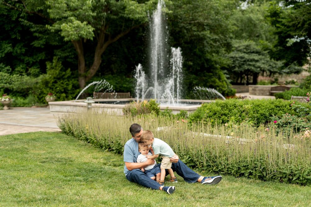 Father and young child sitting together on grass near a beautiful park fountain on a sunny day.