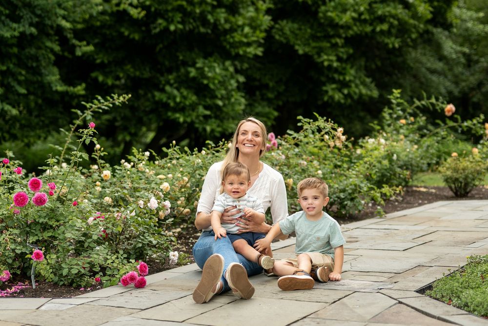 Mother sitting with two young children on a stone path surrounded by roses.