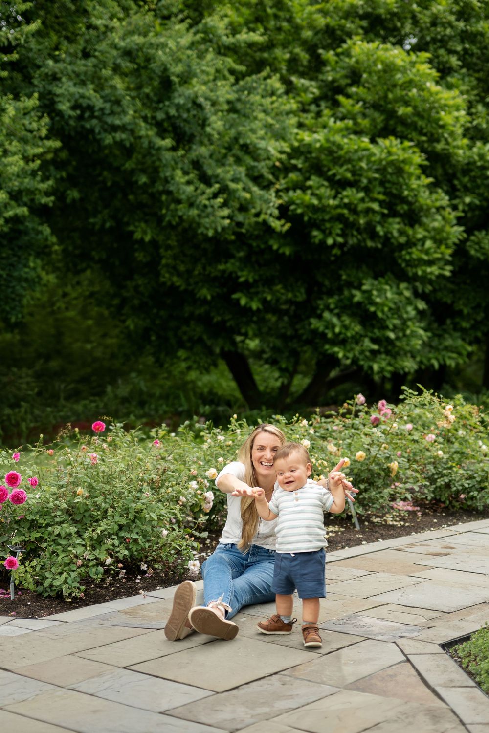 A smiling mother sits on a garden path with her happy toddler among blooming roses and lush green trees.