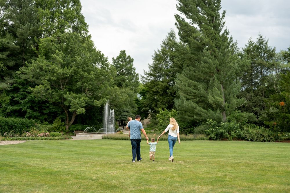 Family of three walking hand-in-hand across green lawn with tall trees and fountain backdrop.
