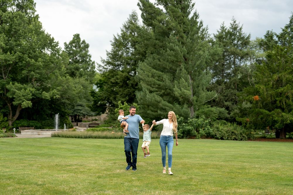 Family of four walking and holding hands on lush green lawn in a park surrounded by tall trees.