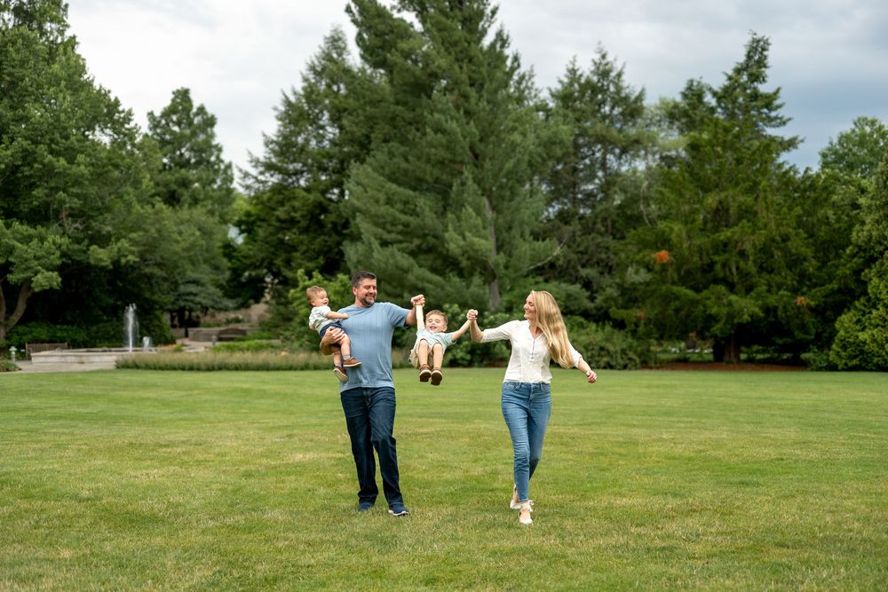 Happy family playing outdoors on green grass, parents lifting children with trees in background.