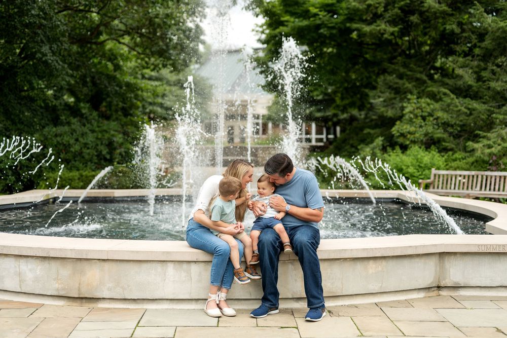 Family of three sitting on fountain edge, parents kissing baby outdoors.