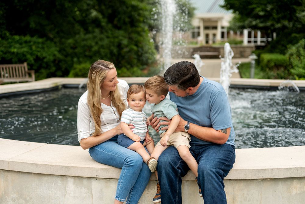 Family with toddler sitting by garden fountain, parents lovingly engaging with child.