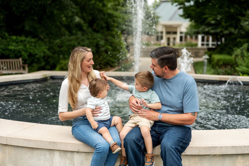 Happy family with two toddlers laughing together by a garden fountain on a sunny day.