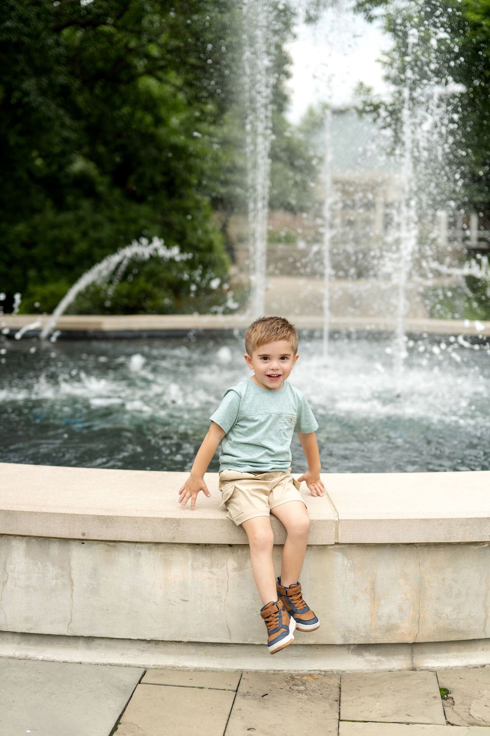 Young boy sitting on fountain edge in mint shirt and khaki shorts, smiling outdoors near water feature.